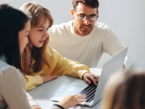 A young girl with light brown hair sits between her mother and father and reviews information on a laptop.