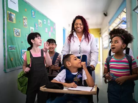 A young Black woman teacher smiles and walks down the hall with four of her elementary school students, while pushing one of them in a wheelchair.
